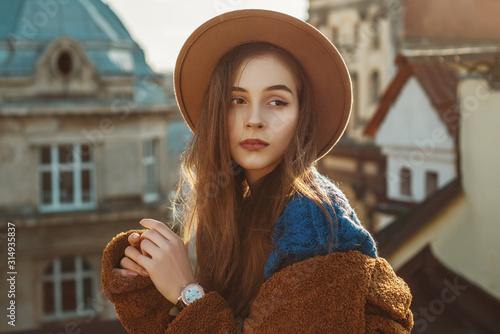 Elegant fashionable brunette woman, model wearing stylish hat, wrist watch, blue sweater, brown faux fur coat, posing in European city. Copy empty space for text