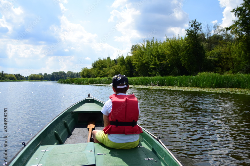 Człowiek w czerwonym kapoku na zielonej łódce płynie rzeką w letni dzień.