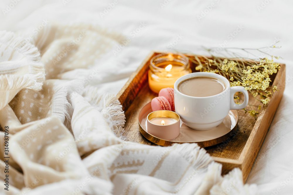 Wooden tray of coffee and candles on bed. White bedding sheets with striped blanket and pillow. Breakfast in bed. Hygge concept. Wooden tray of coffee and candles on bed. White bedding sheets with striped blanket and pillow. Breakfast in bed. Hygge concept.