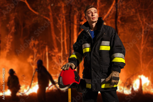 Horizontal view. Firefighter with sad equipment, holds the helmet in his hand, looking up, which stands against the background of a people and forest in flames.