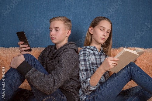Two teenage boy and girl reading together indoor using a phone and a paper book.