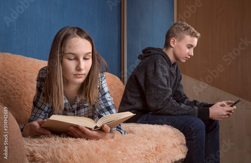 Two teenage boy and girl reading together indoor using a phone and a paper book.