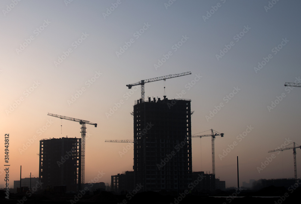 Silhouettes of tower cranes constructing a new residential building at a construction site against sunset background. Renovation program, development, concept of the buildings industry. Silhouettes of tower cranes constructing a new residential building at a construction site against sunset background. Renovation program, development, concept of the buildings industry.