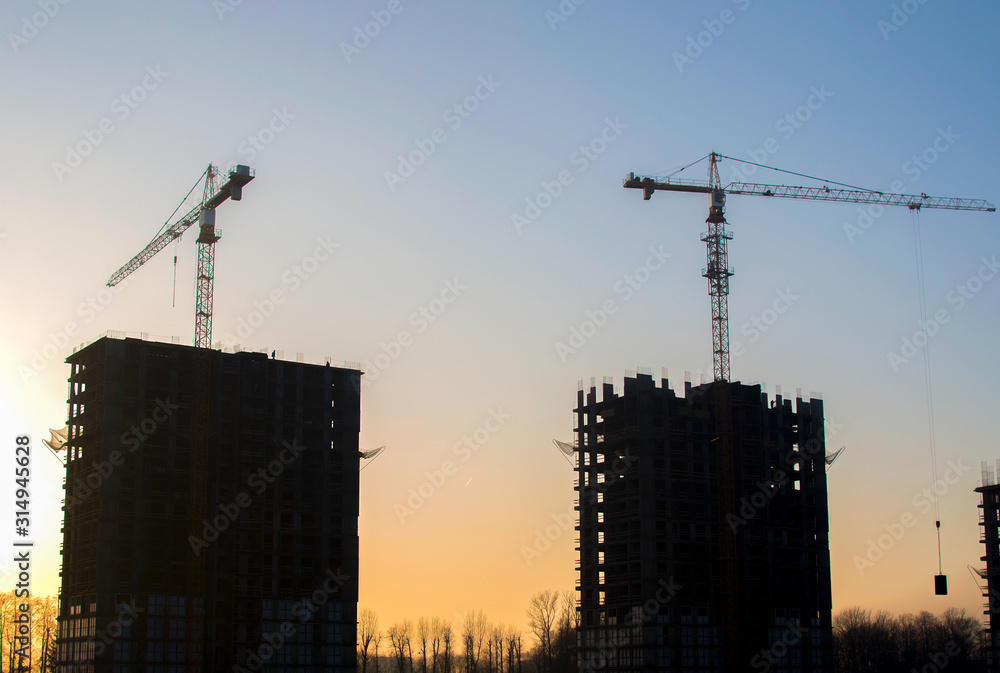 Silhouettes of tower cranes constructing a new residential building at a construction site against sunset background. Renovation program, development, concept of the buildings industry. Silhouettes of tower cranes constructing a new residential building at a construction site against sunset background. Renovation program, development, concept of the buildings industry.