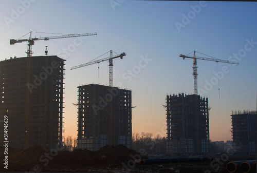 Silhouettes of tower cranes constructing a new residential building at a construction site against sunset background. Renovation program, development, concept of the buildings industry.