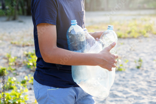 Volunteer boy in blue t shirt picks up dirty plastic bottles in park. Hands in yellow gloves collect garbage, pick up trash in bag. Eco activist cleaning up nature from plastic