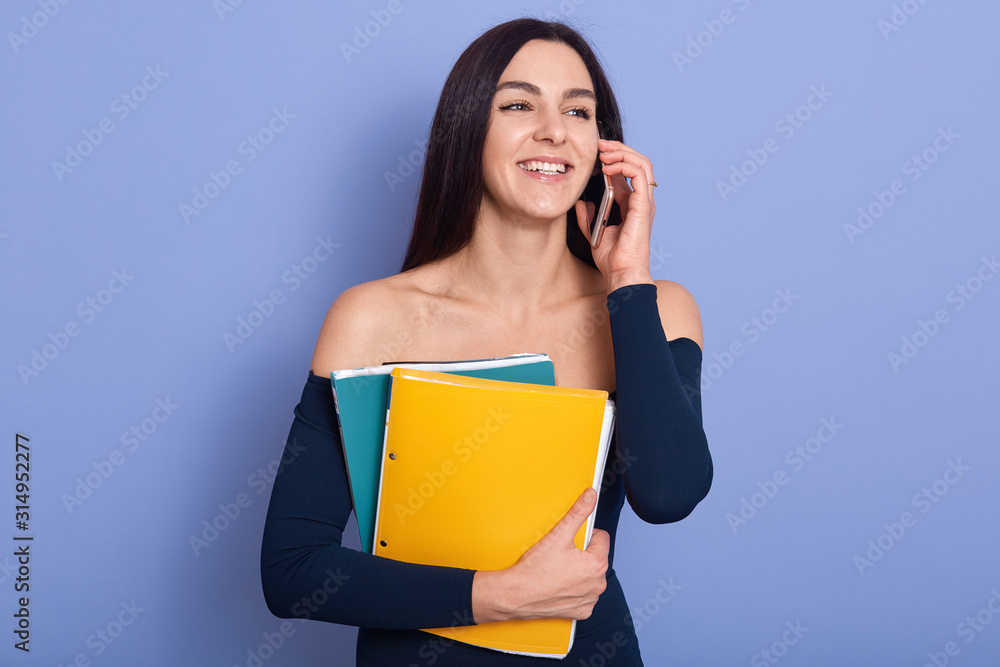 Studio shot of beautiful woman posing over blue background, holding yellow paper folders in hands, looking smiling aside, buisnesswoman has conversation with buisness partner, looks confident. Studio shot of beautiful woman posing over blue background, holding yellow paper folders in hands, looking smiling aside, buisnesswoman has conversation with buisness partner, looks confident.