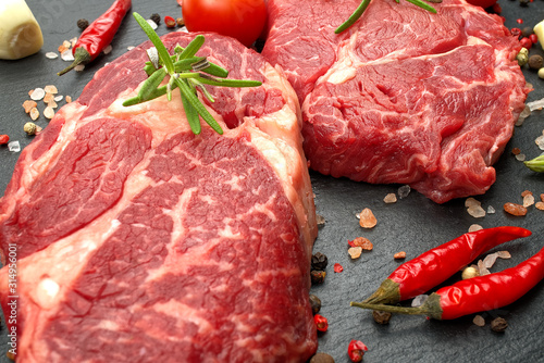 Raw rib eye steak with spices and vegetables. Ingredients for restaurant meal. Fresh meat, salt, rosemary, thyme, chilli, cherry tomatoes, garlic on black stone. Food background.