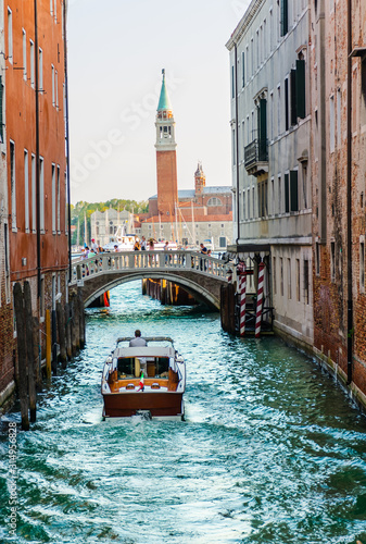 vaporetto amongst tall buildings cruising a venetian canal to the gran canal with a bridge plenty of tourists at the back and beyond the church of Maggiori with its big tower. Vertical photo.