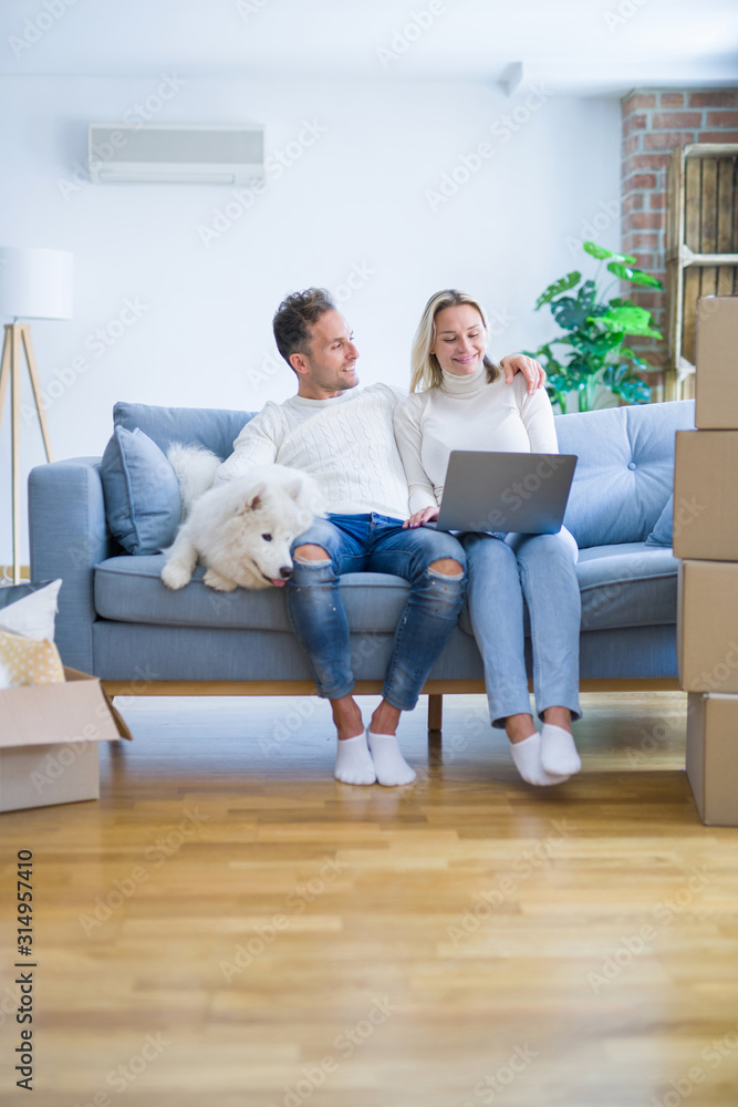 Young beautiful couple with dog sitting on the sofa using laptop at new home around cardboard boxes Young beautiful couple with dog sitting on the sofa using laptop at new home around cardboard boxes