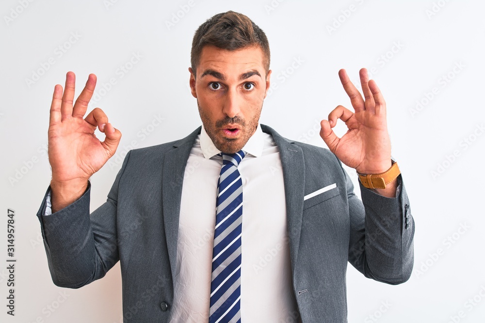 Young handsome business man wearing suit and tie over isolated background looking surprised and shocked doing ok approval symbol with fingers. Crazy expression - obrazy, fototapety, plakaty 