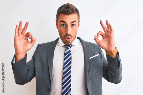Young handsome business man wearing suit and tie over isolated background looking surprised and shocked doing ok approval symbol with fingers. Crazy expression