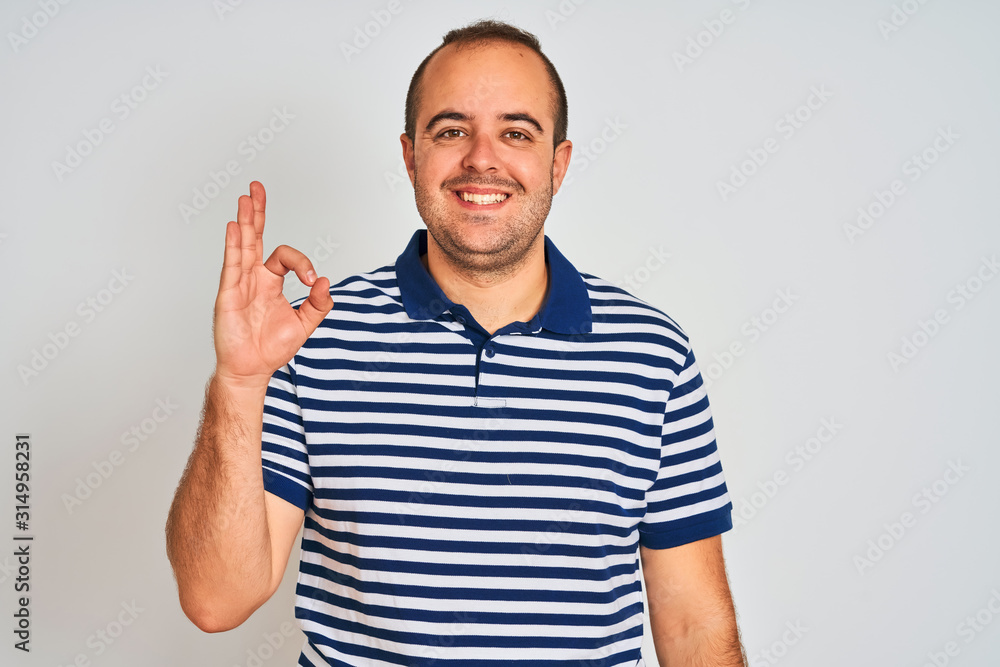 Naklejka premium Young man wearing casual striped polo standing over isolated white background smiling positive doing ok sign with hand and fingers. Successful expression.