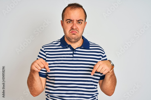 Young man wearing casual striped polo standing over isolated white background Pointing down looking sad and upset, indicating direction with fingers, unhappy and depressed.