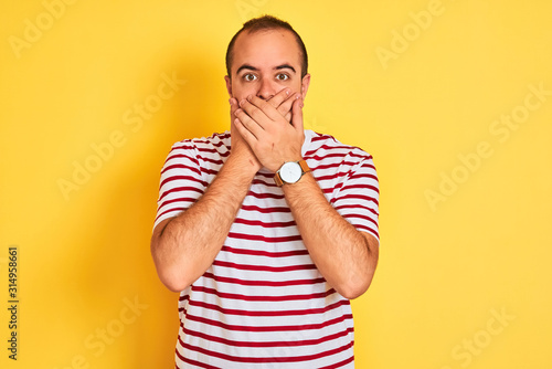 Young man wearing casual striped t-shirt standing over isolated yellow background shocked covering mouth with hands for mistake. Secret concept.