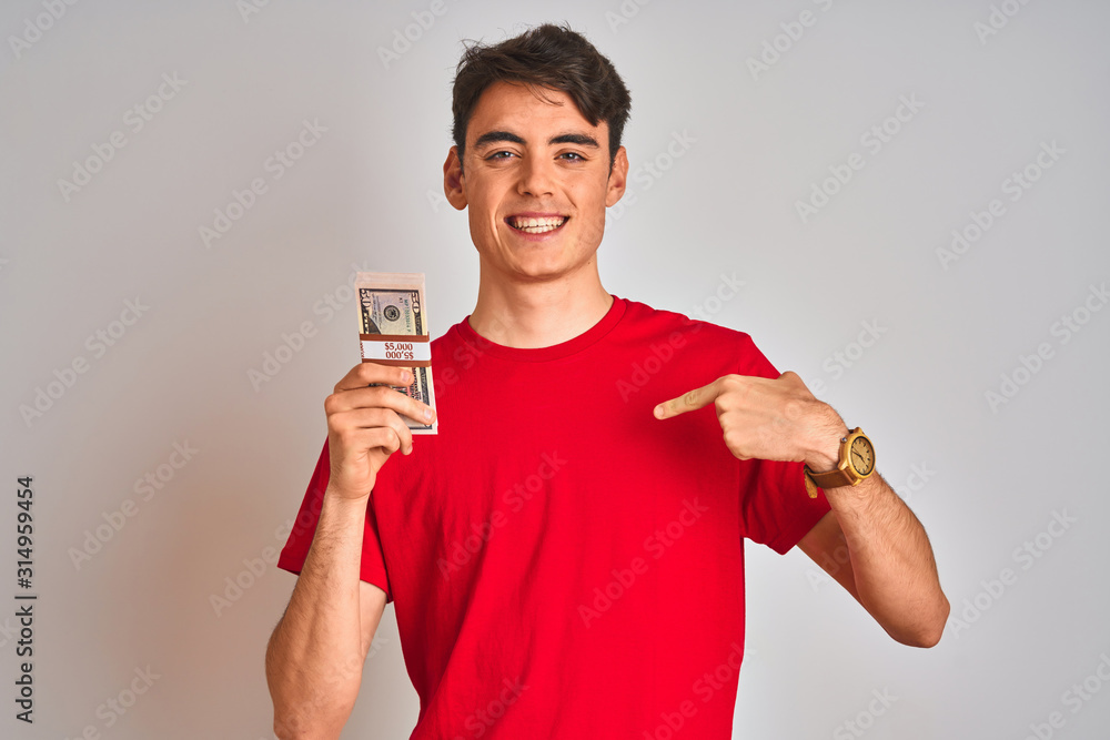 Teenager boy holding a bunch of dollars bank notes over isolated background with surprise face pointing finger to himself Teenager boy holding a bunch of dollars bank notes over isolated background with surprise face pointing finger to himself
