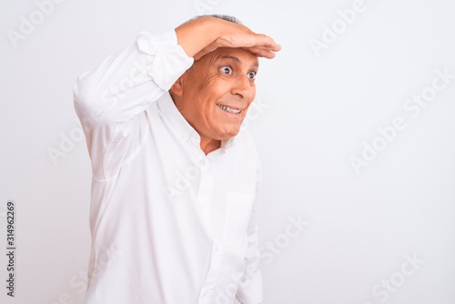 Senior grey-haired man wearing elegant shirt standing over isolated white background very happy and smiling looking far away with hand over head. Searching concept.