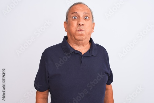 Senior grey-haired man wearing black casual polo standing over isolated white background puffing cheeks with funny face. Mouth inflated with air, crazy expression.