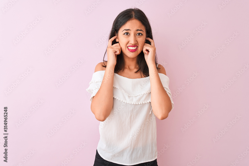 Beautiful chinese woman wearing white t-shirt standing over isolated pink background covering ears with fingers with annoyed expression for the noise of loud music. Deaf concept. Beautiful chinese woman wearing white t-shirt standing over isolated pink background covering ears with fingers with annoyed expression for the noise of loud music. Deaf concept.