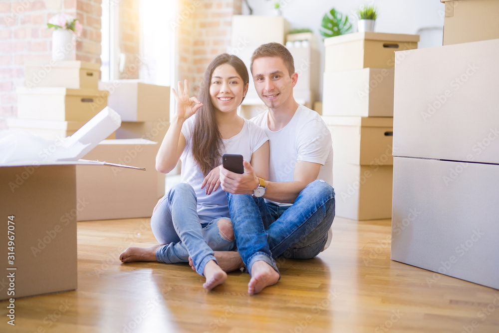 Beautiful couple sitting on the floor using smartphone at new home around cardboard boxes doing ok sign with fingers, excellent symbol Beautiful couple sitting on the floor using smartphone at new home around cardboard boxes doing ok sign with fingers, excellent symbol