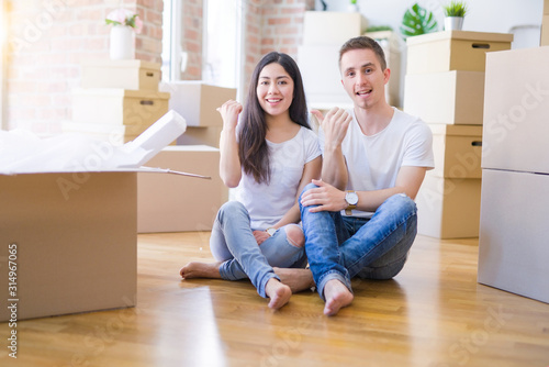Young beautiful couple sitting on the floor at new home around cardboard boxes pointing and showing with thumb up to the side with happy face smiling