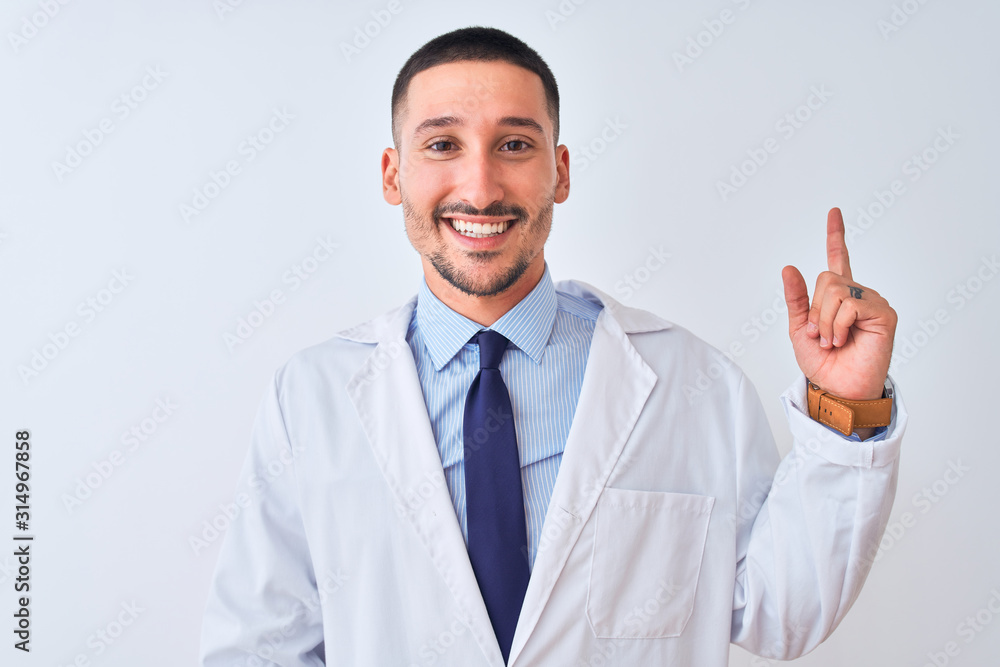 Young doctor man wearing white coat over isolated background with a big smile on face, pointing with hand and finger to the side looking at the camera. Young doctor man wearing white coat over isolated background with a big smile on face, pointing with hand and finger to the side looking at the camera.