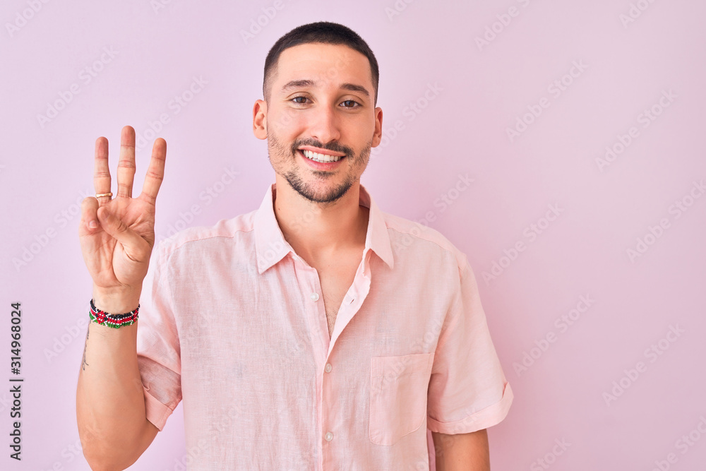 Young handsome man wearing pink shirt standing over isolated background showing and pointing up with fingers number three while smiling confident and happy. Young handsome man wearing pink shirt standing over isolated background showing and pointing up with fingers number three while smiling confident and happy.