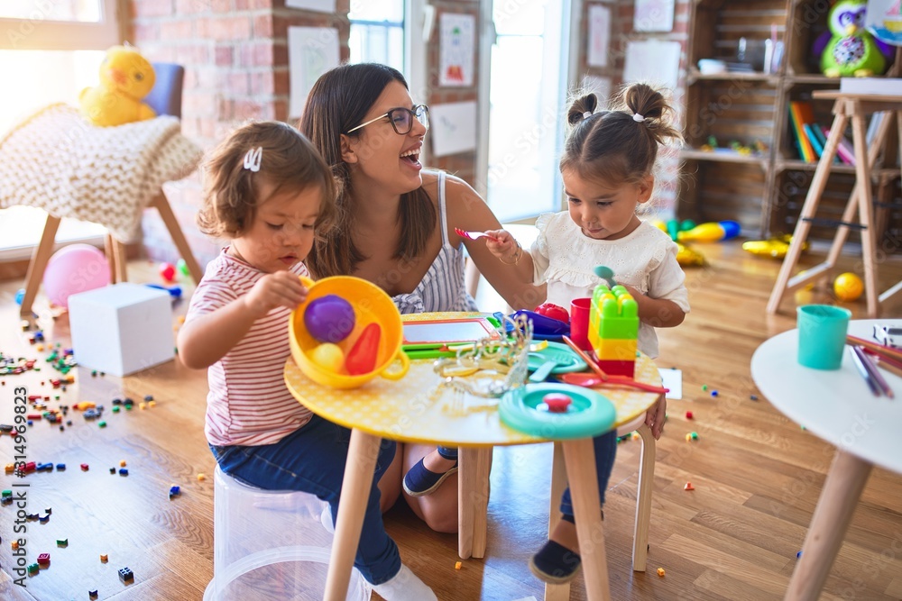 Young beautiful teacher and toddlers playing meals using plastic food and cutlery toy at kindergarten - obrazy, fototapety, plakaty 