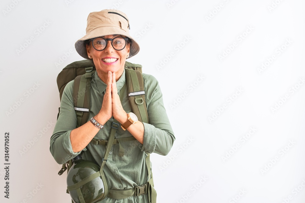 Middle age hiker woman wearing backpack canteen hat glasses over isolated white background praying with hands together asking for forgiveness smiling confident. Middle age hiker woman wearing backpack canteen hat glasses over isolated white background praying with hands together asking for forgiveness smiling confident.