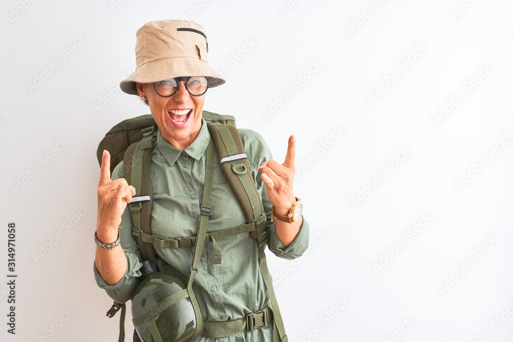 Middle age hiker woman wearing backpack canteen hat glasses over isolated white background shouting with crazy expression doing rock symbol with hands up. Music star. Heavy concept. Middle age hiker woman wearing backpack canteen hat glasses over isolated white background shouting with crazy expression doing rock symbol with hands up. Music star. Heavy concept.
