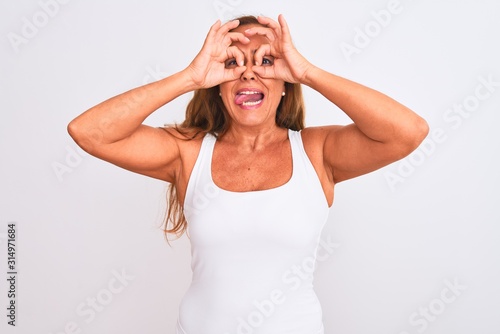 Middle age mature woman standing over white isolated background doing ok gesture like binoculars sticking tongue out, eyes looking through fingers. Crazy expression.