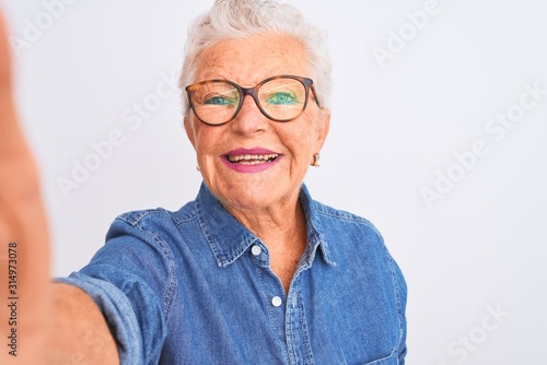 Grey-haired woman wearing denim shirt and glasses make selfie over isolated white background with a happy face standing and smiling with a confident smile showing teeth