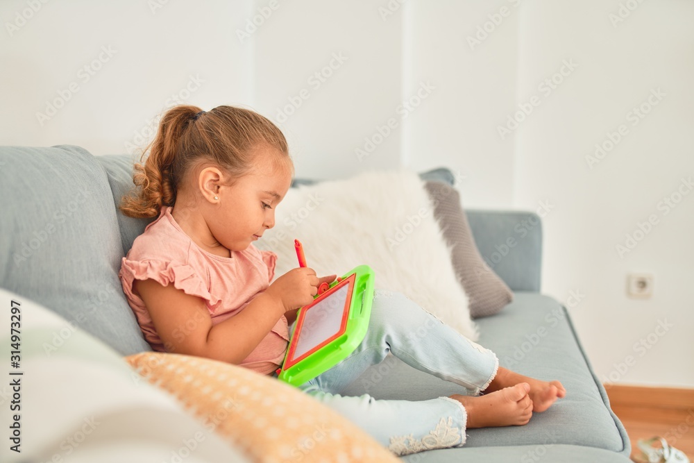 Beautiful blond toddler girl sitting on the sofa drawing using digital board at home Beautiful blond toddler girl sitting on the sofa drawing using digital board at home