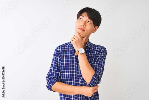 Young chinese man wearing casual blue shirt standing over isolated white background with hand on chin thinking about question, pensive expression. Smiling with thoughtful face. Doubt concept.