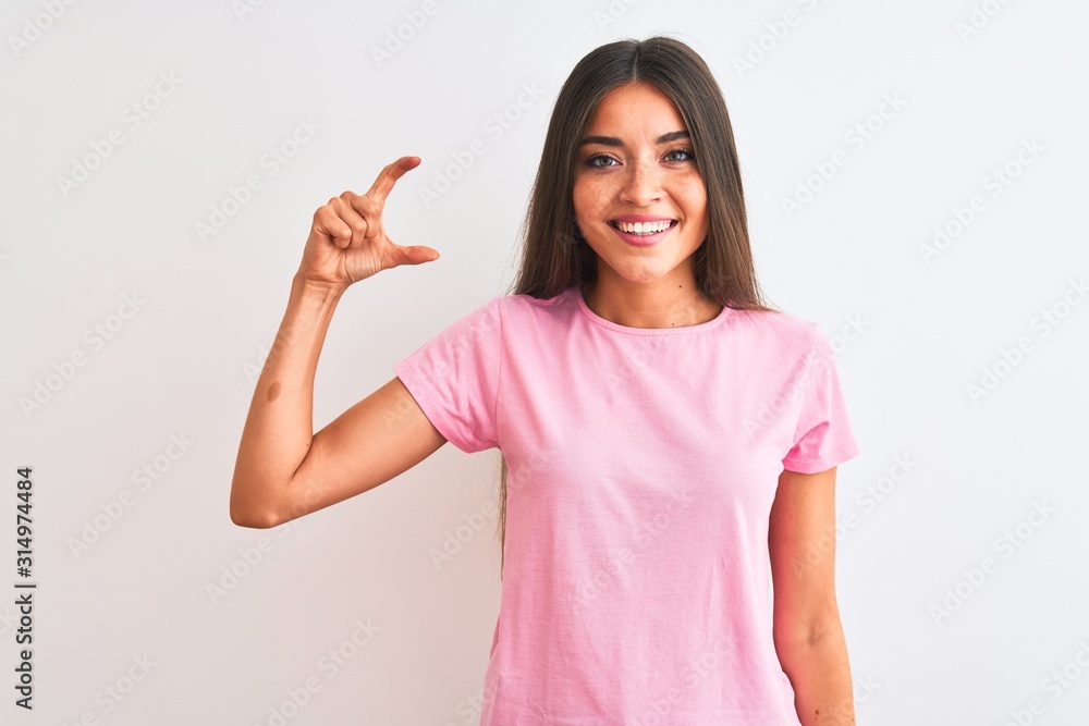 Young beautiful woman wearing pink casual t-shirt standing over isolated white background smiling and confident gesturing with hand doing small size sign with fingers looking and the camera. Measure Young beautiful woman wearing pink casual t-shirt standing over isolated white background smiling and confident gesturing with hand doing small size sign with fingers looking and the camera. Measure