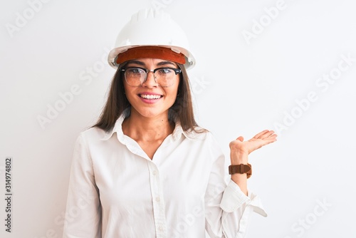 Young beautiful architect woman wearing helmet and glasses over isolated white background smiling cheerful presenting and pointing with palm of hand looking at the camera.