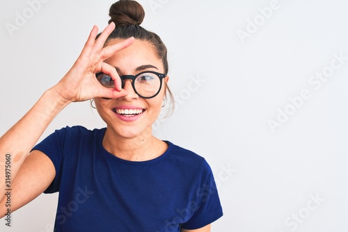 Beautiful woman with bun wearing blue t-shirt and glasses over isolated white background with happy face smiling doing ok sign with hand on eye looking through fingers