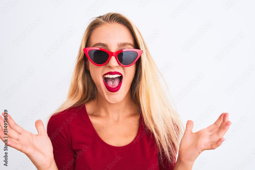 Young beautiful woman wearing red t-shirt and sunglasses over isolated white background celebrating crazy and amazed for success with arms raised and open eyes screaming excited. Winner concept Young beautiful woman wearing red t-shirt and sunglasses over isolated white background celebrating crazy and amazed for success with arms raised and open eyes screaming excited. Winner concept