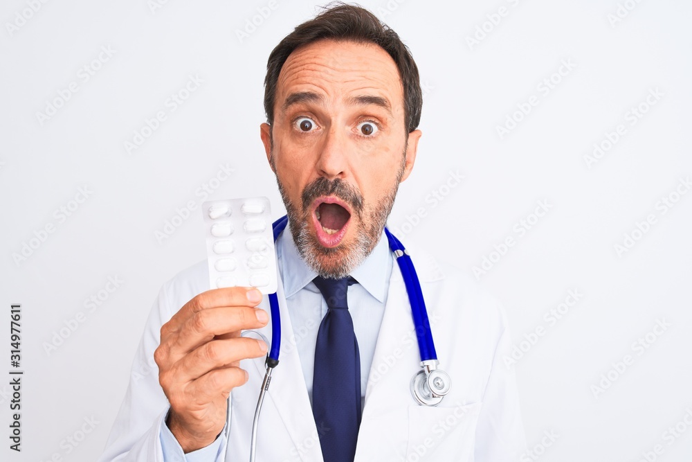 Middle age doctor man holding pills standing over isolated white background scared in shock with a surprise face, afraid and excited with fear expression Middle age doctor man holding pills standing over isolated white background scared in shock with a surprise face, afraid and excited with fear expression