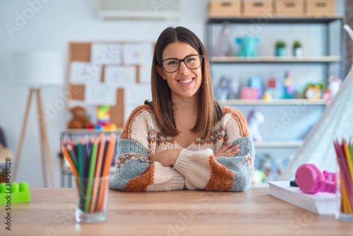 Young beautiful teacher woman wearing sweater and glasses sitting on desk at kindergarten happy face smiling with crossed arms looking at the camera. Positive person. photo