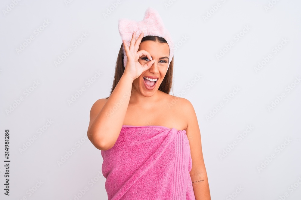 Beautiful young woman wearing pink shower towel and beauty headband over white background doing ok gesture with hand smiling, eye looking through fingers with happy face. Beautiful young woman wearing pink shower towel and beauty headband over white background doing ok gesture with hand smiling, eye looking through fingers with happy face.