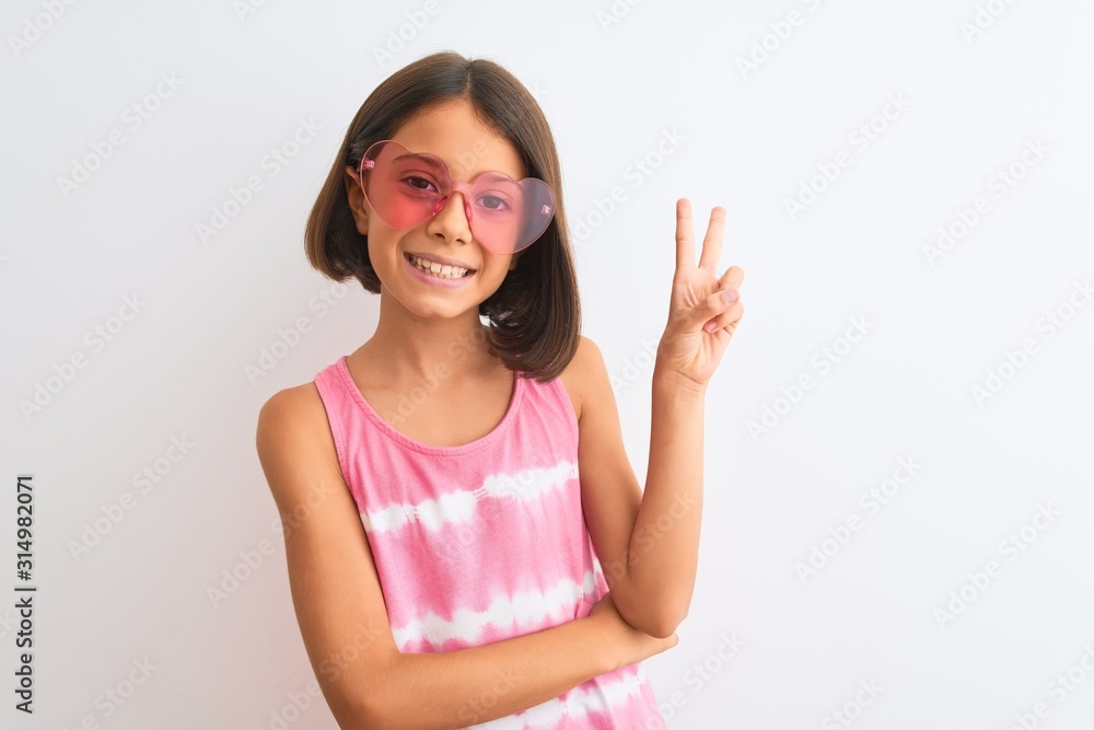 Young beautiful child girl wearing pink t-shirt and sunglasses over isolated white background smiling with happy face winking at the camera doing victory sign. Number two. Young beautiful child girl wearing pink t-shirt and sunglasses over isolated white background smiling with happy face winking at the camera doing victory sign. Number two.