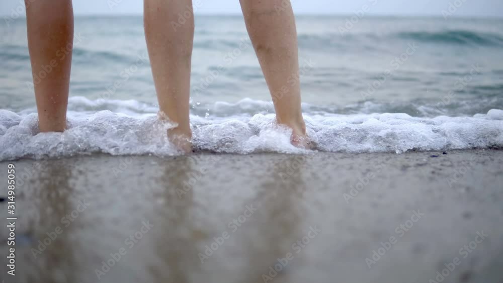 Two pairs of feet stand on the sand and wait for the wave to come. wave on the sandy beach, cinematic Two pairs of feet stand on the sand and wait for the wave to come. wave on the sandy beach, cinematic