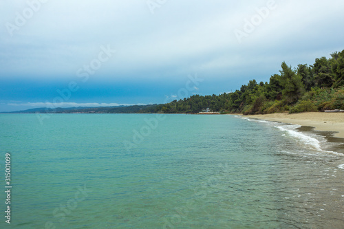 Beautiful coastal line of Greece. Horizontal color photography of clear blue sea water, cloudy sky, green hills and sandy beach.