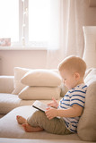 Cute, beautiful toddler boy sitting on a sofa at home and playing educational games on a tablet. Educational games and early childhood development. Lifestyle concept