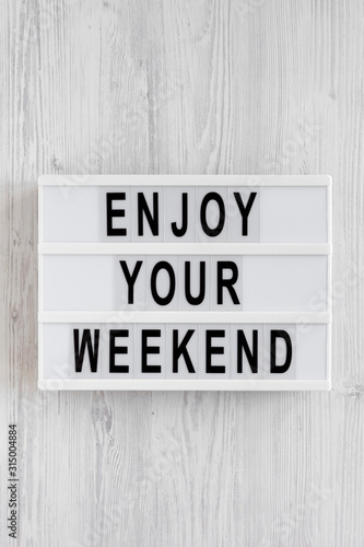 'Enjoy your weekend' words on a lightbox on a white wooden surface, top view. Overhead, from above, flat lay. Space for text.