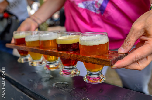 Craft beer tasting: five glasses with beers of different colors and flavors. A wooden cup holder holds the glasses in a row. Two Caucasian woman's hands hold him up.