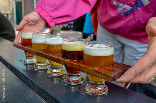 Craft beer tasting: five glasses with beers of different colors and flavors. A wooden cup holder holds the glasses in a row. Two Caucasian woman's hands hold him up.