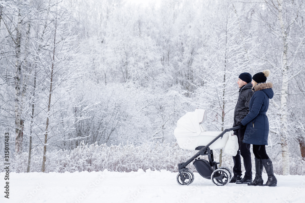 Happy young family pushing white baby stroller and walking at nature park. Snow covered trees and bushes. Spending time with infant in beautiful winter day. Enjoying peaceful stroll. Side view. Happy young family pushing white baby stroller and walking at nature park. Snow covered trees and bushes. Spending time with infant in beautiful winter day. Enjoying peaceful stroll. Side view.