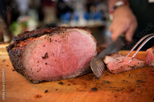 Chef slicing a prime rib roast, rare, with a knife and a meat fork on a wooden cutting board, in a restaurant